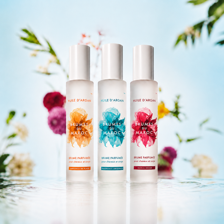 Brumes parfumées Moroccanoil pour cheveux et corps avec les fragrances Ambiance de Plage, Fragrance Originale et Dahlia Rouge dans des flacons givrés blancs sur fond floral lumineux bleu pastel.