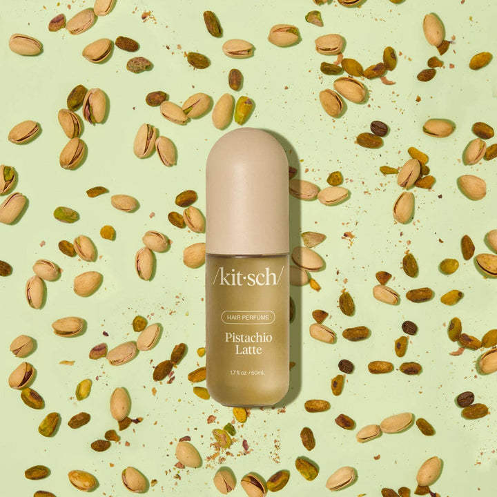 Parfum pour cheveux KITSCH Pistachio Latte 50 ml présenté sur un fond vert entouré de pistaches. Flacon beige au design minimaliste évoquant une fragrance gourmande à la pistache, noix de coco et café.