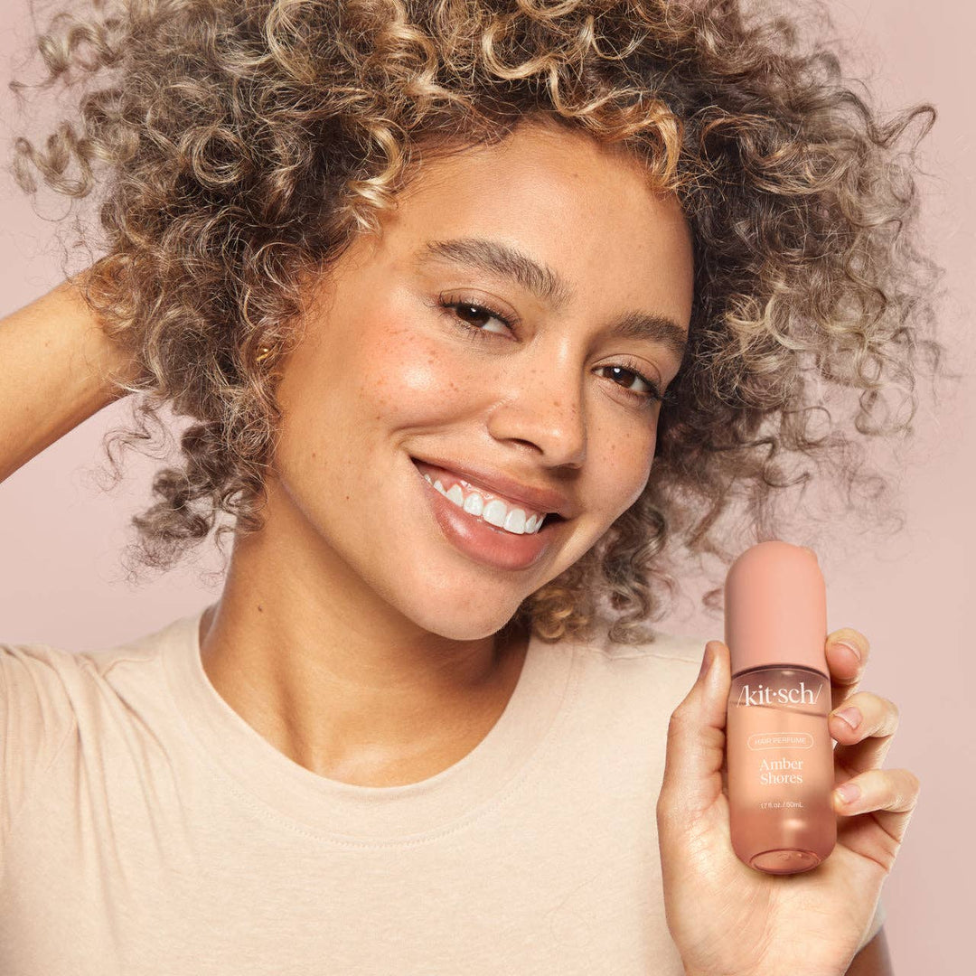 Femme souriante tenant le parfum pour cheveux KITSCH Amber Shores 50 ml, démonstration du produit pour des cheveux délicatement parfumés et rafraîchis.