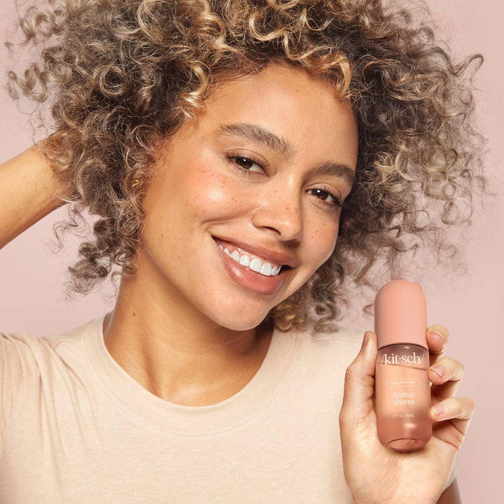 Femme souriante tenant le parfum pour cheveux KITSCH Amber Shores 50 ml, démonstration du produit pour des cheveux délicatement parfumés et rafraîchis.