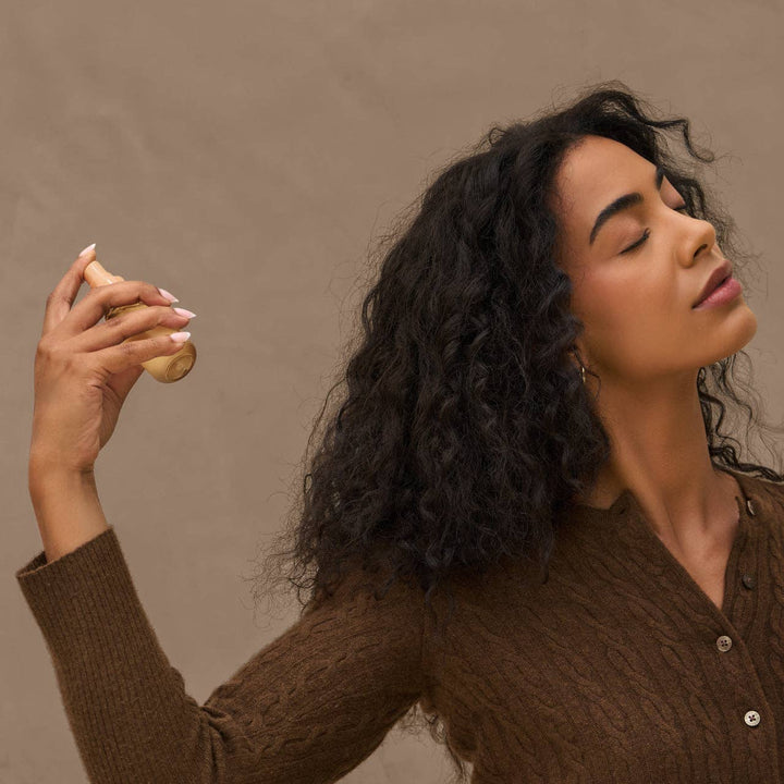 Femme aux cheveux bouclés vaporisant le parfum pour cheveux KITSCH Vanille Rustique. Brume légère parfumée pour rafraîchir les cheveux entre deux shampoings.