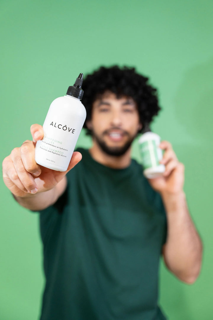Homme aux cheveux bouclés tenant une bouteille de shampooing Alcôve devant un fond vert, mettant en valeur le produit pour cheveux texturés.