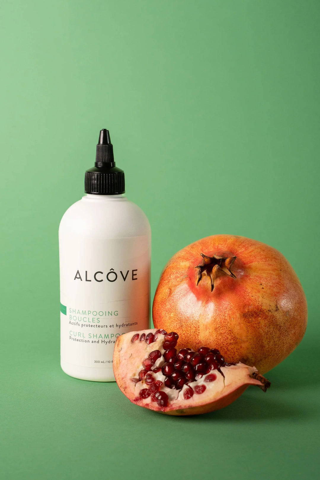 Shampooing boucles Alcôve placé à côté d’une grenade entière et d’une moitié ouverte, illustrant les ingrédients naturels et l'hydratation du produit.