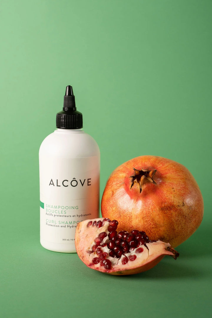Shampooing boucles Alcôve placé à côté d’une grenade entière et d’une moitié ouverte, illustrant les ingrédients naturels et l'hydratation du produit.