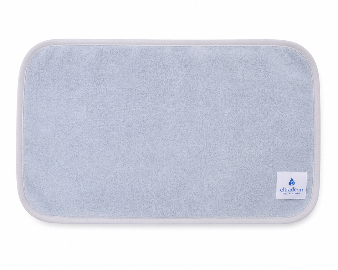 Serviette nettoyante pour le visage Eltraderm en microfibres gris pâle avec bordure assortie et étiquette de marque visible, présentée à plat sur fond blanc. Accessoire réutilisable et lavable conçu pour nettoyer la peau en douceur.
