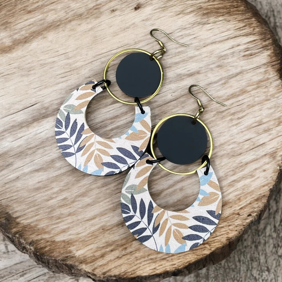 Paire de boucles d’oreilles pendantes avec pendentifs en bois imprimé de motifs feuillagés aux tons neutres, anneau doré et pastille noire, sur fond de bois naturel.