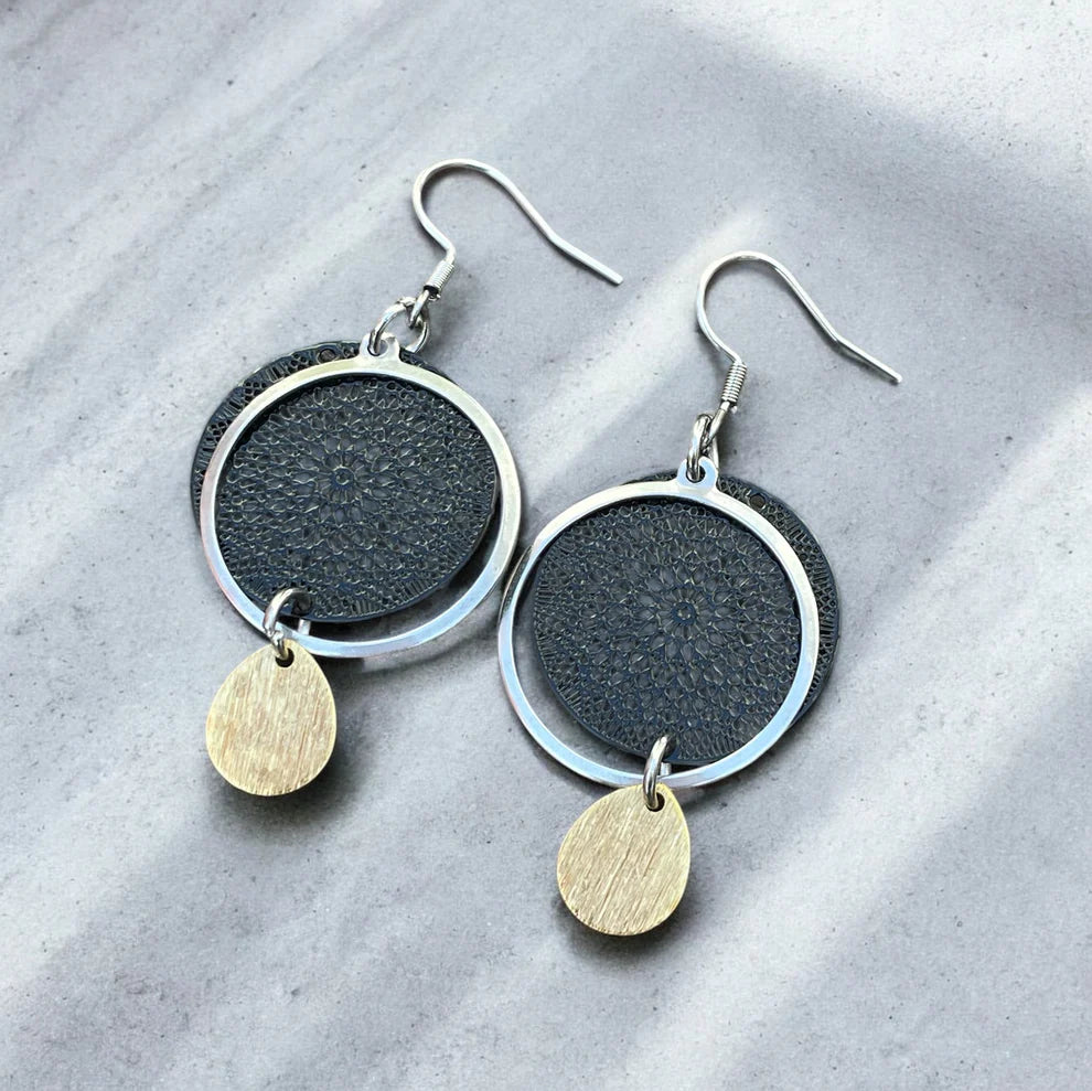 Paire de boucles d’oreilles artisanales composées d’un cercle noir texturé, d’un anneau en acier inoxydable argenté et d’un pendentif goutte en laiton doré, posées sur un fond gris marbré.