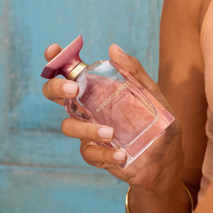 Main tenant un flacon de parfum Moroccanoil Lumière du Jour eau de parfum, bouteille rose translucide avec capuchon métallique rosé.