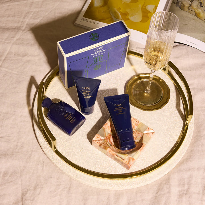 Produits Oribe Supershine formats voyage posés sur un plateau décoratif avec coupe de champagne et coffret découverte bleu dans une ambiance élégante.