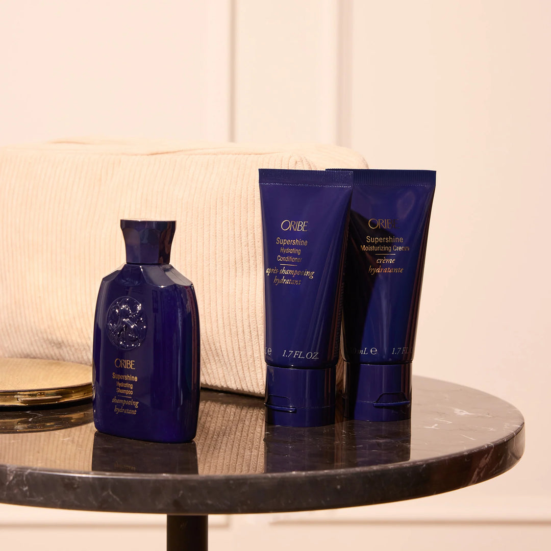 Shampooing hydratant et soins Oribe Supershine formats voyage présentés sur une table en marbre avec trousse beige en arrière-plan.