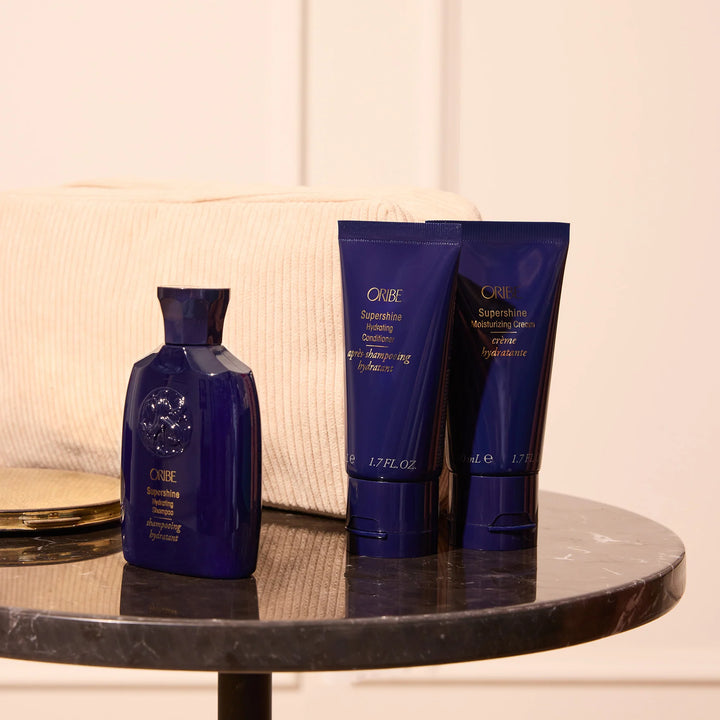 Shampooing hydratant et soins Oribe Supershine formats voyage présentés sur une table en marbre avec trousse beige en arrière-plan.