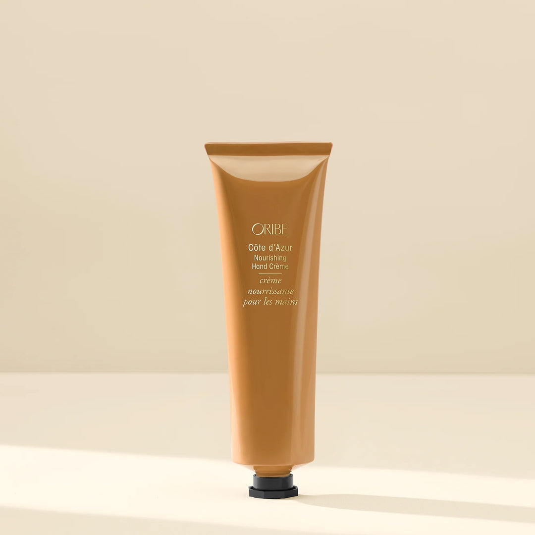 Crème mains nourrissante Oribe Côte d’Azur en tube beige doré au fini brillant avec bouchon noir, présentée sur fond neutre minimaliste, style luxe soin hydratant pour les mains.