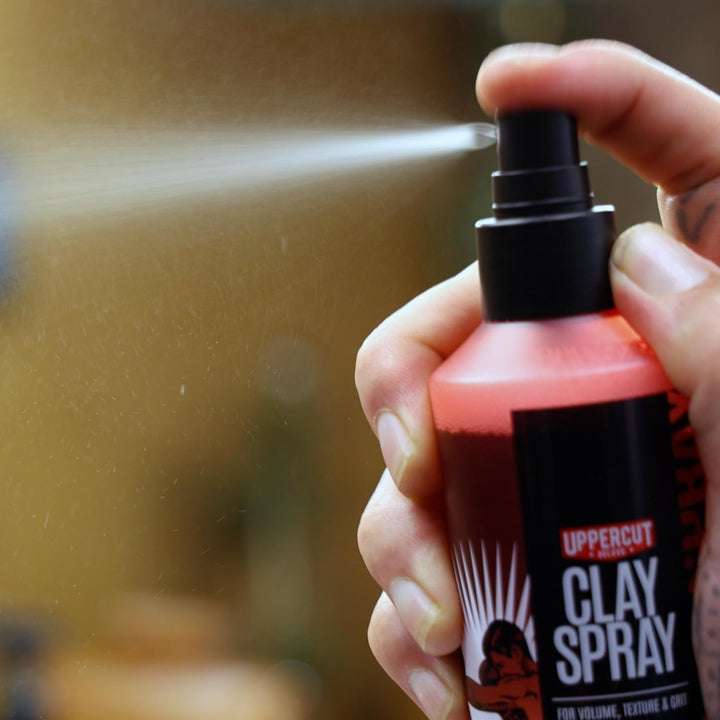 Main vaporisant spray coiffant Uppercut Clay Spray sur fond flou, jet de produit visible sortant du pulvérisateur noir, flacon rouge orangé en gros plan