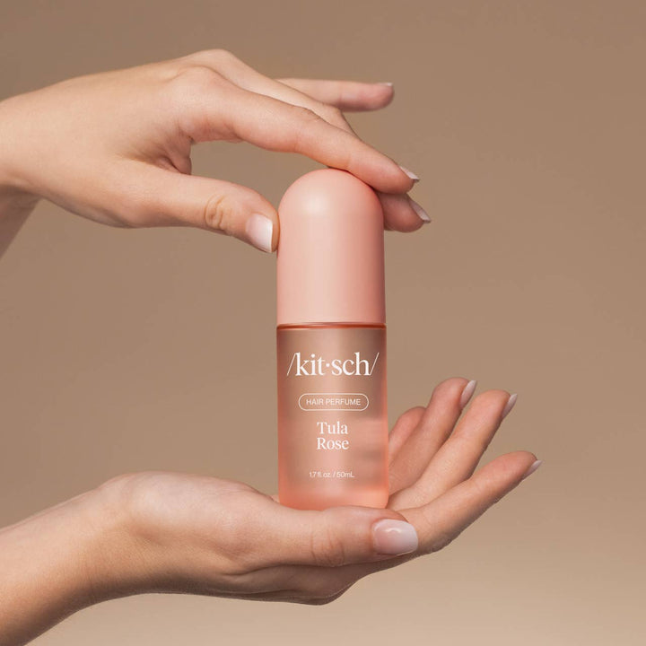 Parfum pour cheveux KITSCH Tula Rose 50 ml tenu entre deux mains, flacon rose givré avec capuchon rose mat. Parfum capillaire floral à la rose, freesia et vanille sur fond beige.