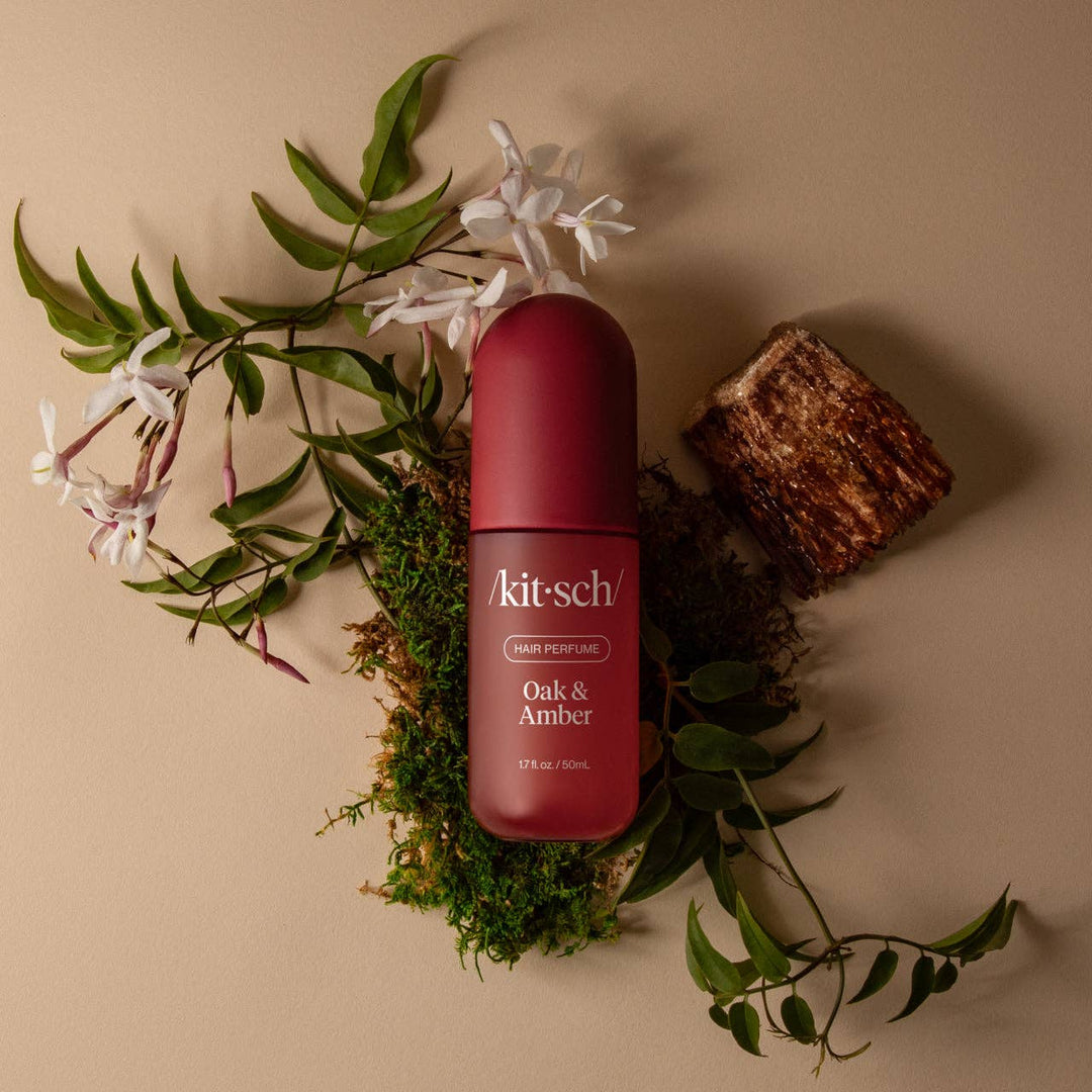 Parfum pour cheveux KITSCH Chêne & Ambre 50 ml présenté sur une pièce de bois avec mousse, fleurs blanches et épices, illustrant sa fragrance boisée et ambrée.