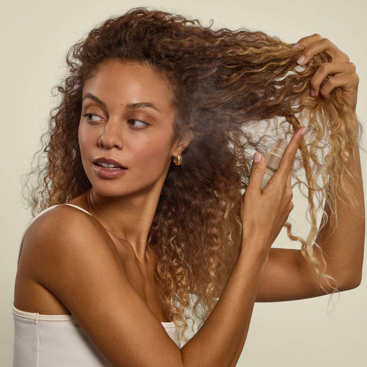 Femme vaporisant le parfum pour cheveux KITSCH Pistachio Latte sur ses cheveux bouclés. Brume légère idéale pour rafraîchir les cheveux entre deux shampoings avec une fragrance gourmande.