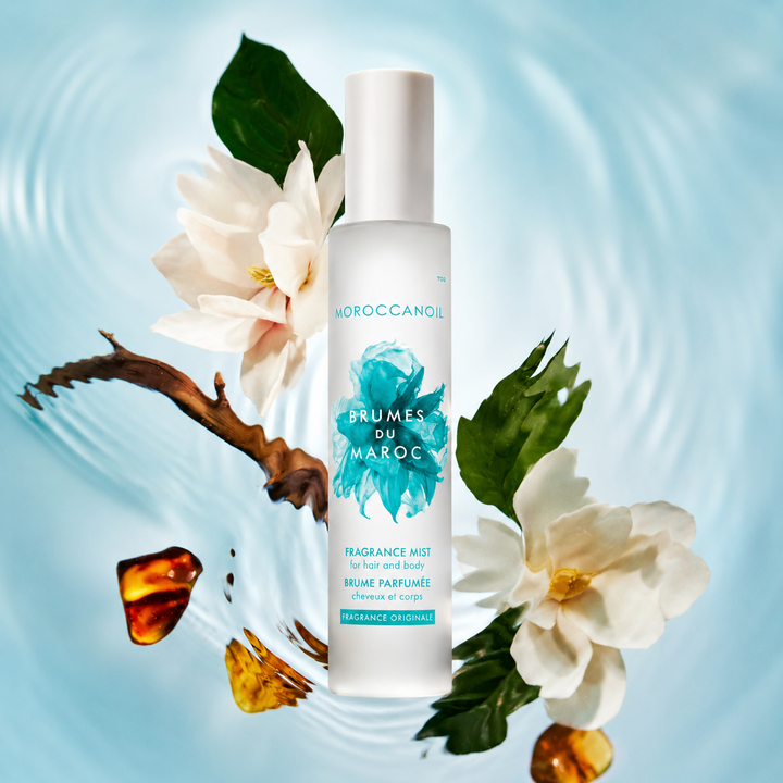 Flacon grand format Moroccanoil Brumes du Maroc Fragrance Originale avec fleurs blanches, feuilles vertes et accents ambrés sur fond bleu clair effet ondulation d’eau.