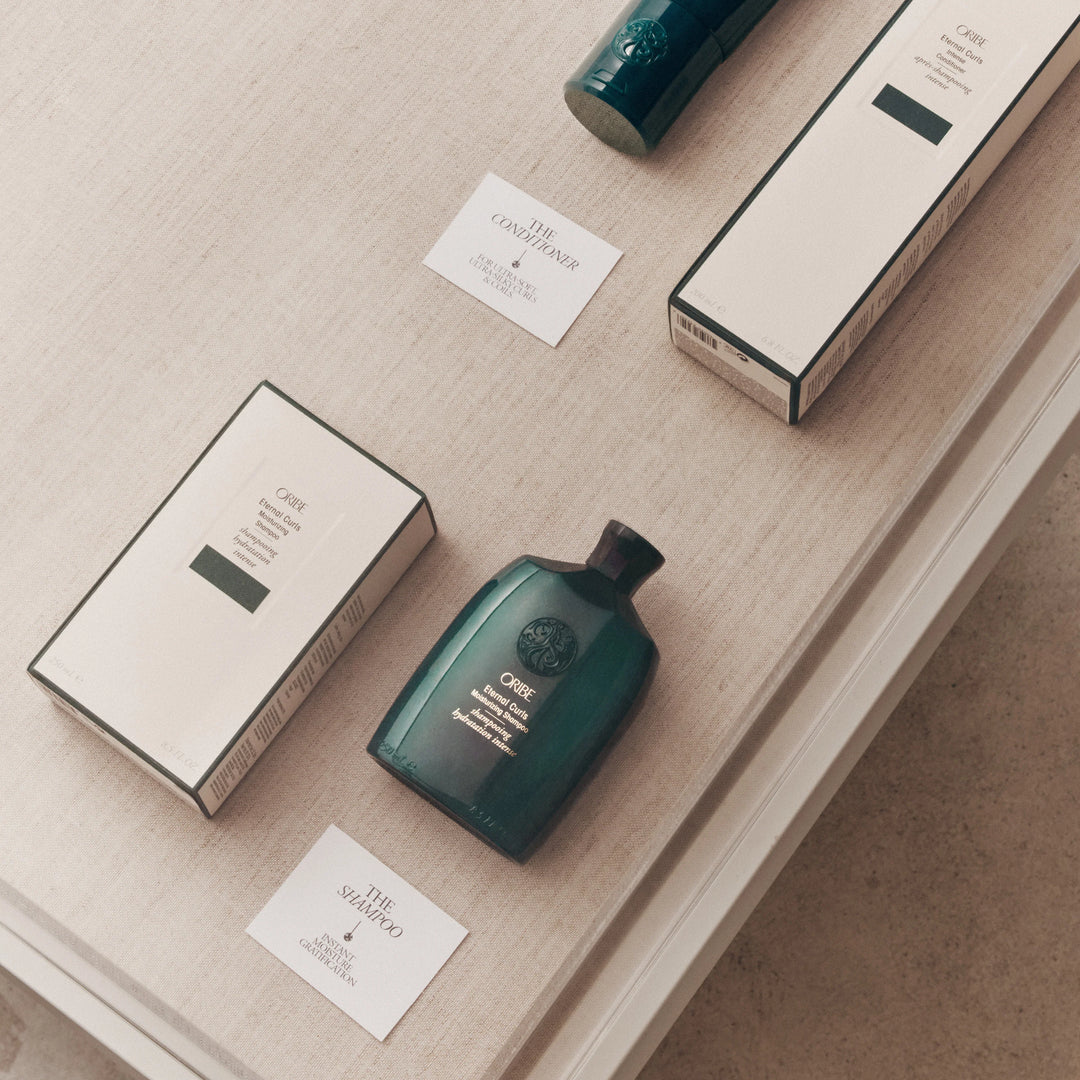 Shampooing et revitalisant Oribe Eternal Curls avec emballages présentés sur une table textile dans une mise en scène élégante de la collection pour cheveux bouclés.