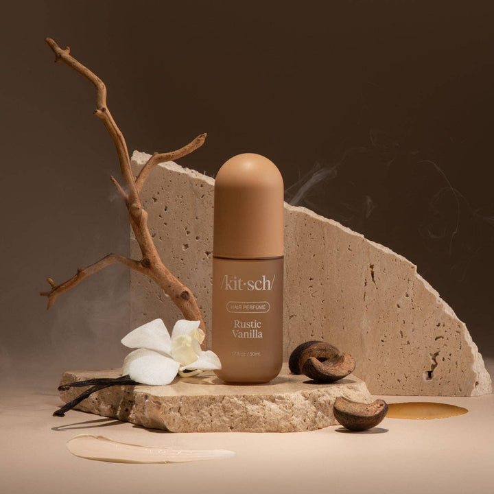 Parfum pour cheveux KITSCH Vanille Rustique 50 ml présenté sur une pierre avec fleur blanche, gousse de vanille, bois et noix. Flacon brun givré sur fond chaleureux évoquant ses notes boisées.