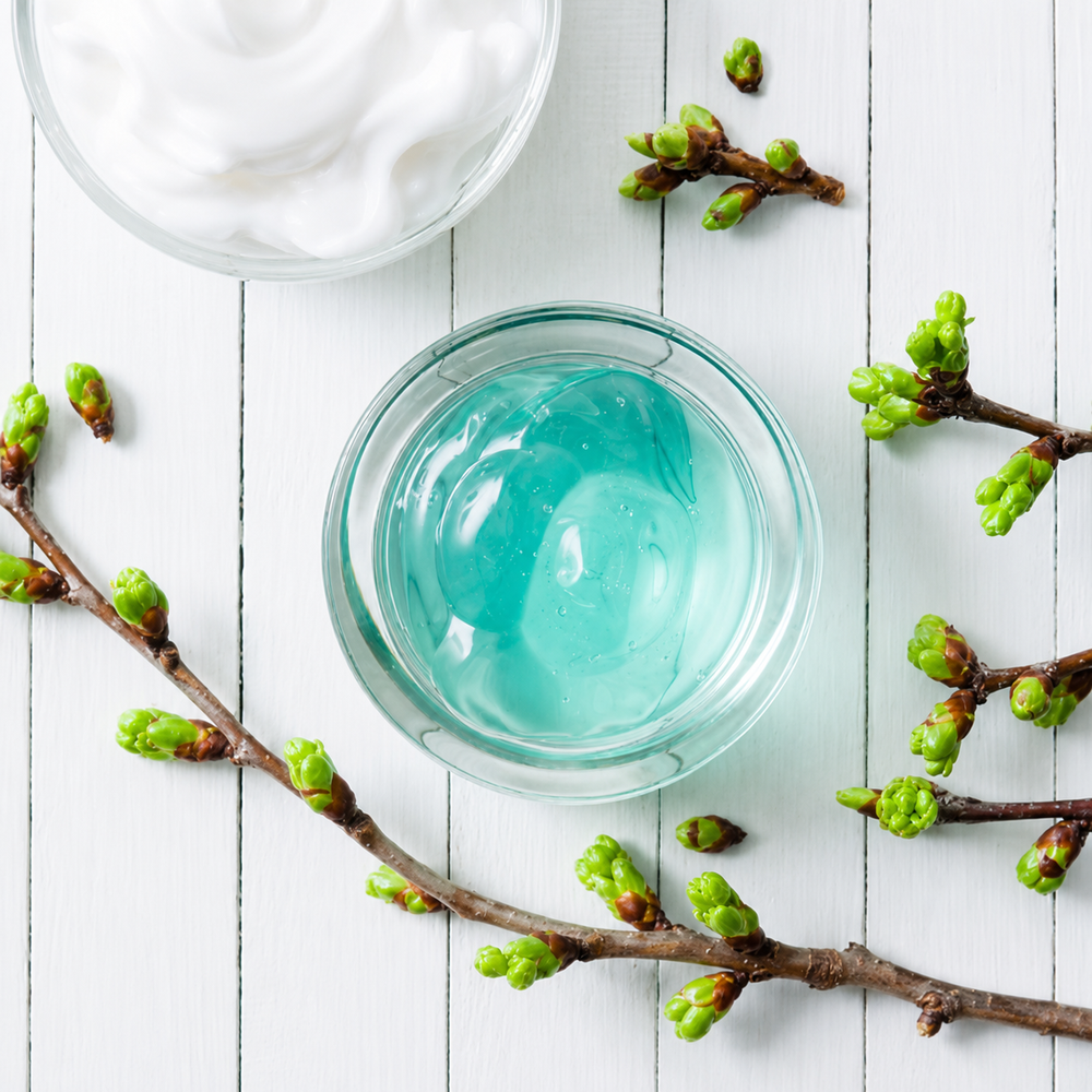 Vue de dessus d’un gel hydratant bleu translucide dans un pot en verre transparent, accompagné d’une crème blanche et de branches aux jeunes bourgeons verts sur fond de bois blanc