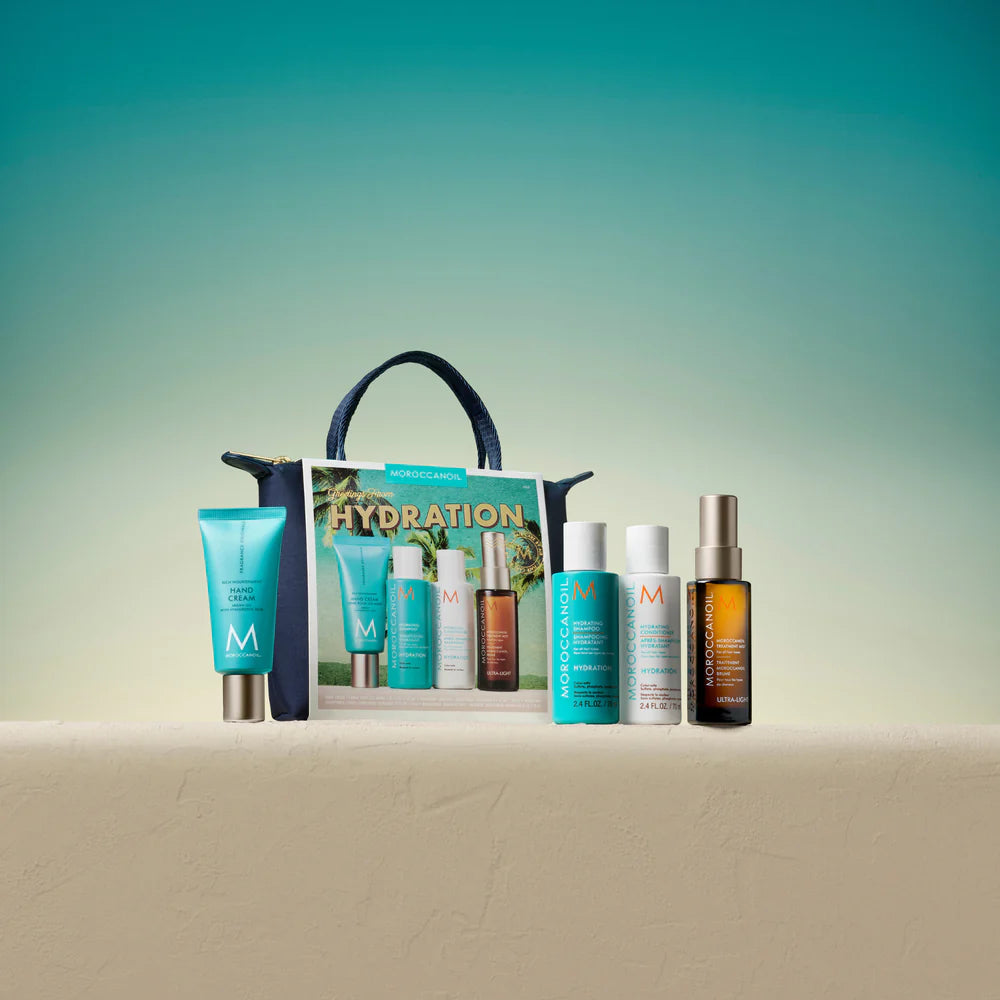 Coffret Moroccanoil Hydration dans une pochette bleu marin avec crème pour les mains, shampoing hydratant, revitalisant et traitement Moroccanoil alignés sur une surface beige devant un fond dégradé vert et turquoise.