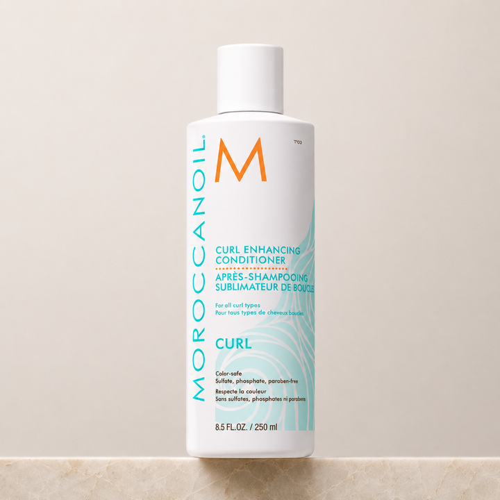 Après-shampoing Sublimateur de Boucles Moroccanoil 250 ml en bouteille blanche avec bouchon blanc, étiquette turquoise, présenté sur un comptoir de pierre beige avec fond neutre.
