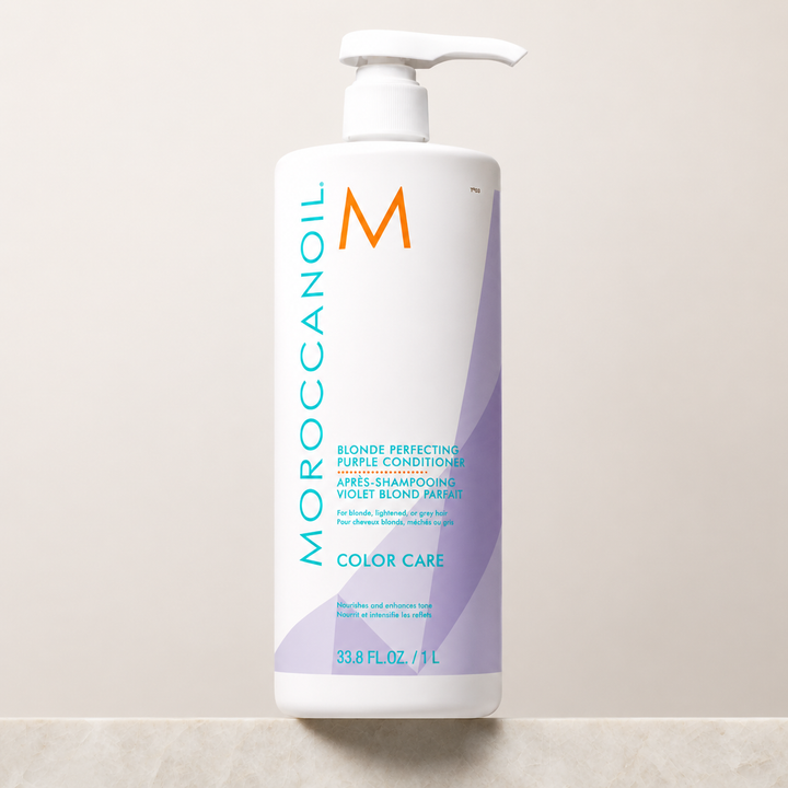 Après-shampooing Violet Blond Parfait Moroccanoil 1 L avec pompe blanche, bouteille blanche ornée de détails violets et turquoise, photographiée sur un comptoir en pierre avec fond beige dans un studio professionnel.